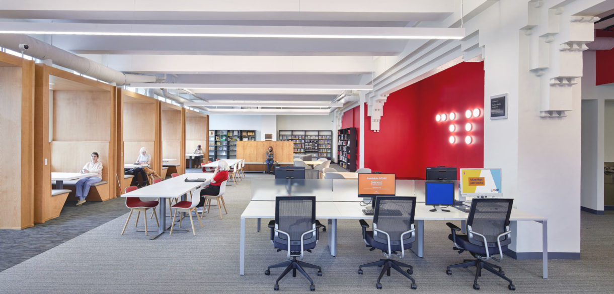 A bank of deep milled-wood booths occupy run along the exterior wall and a bold, red wall with braille supergraphics runs along the facing wall. Between there are tables, chairs, and computers. Books line the back wall.